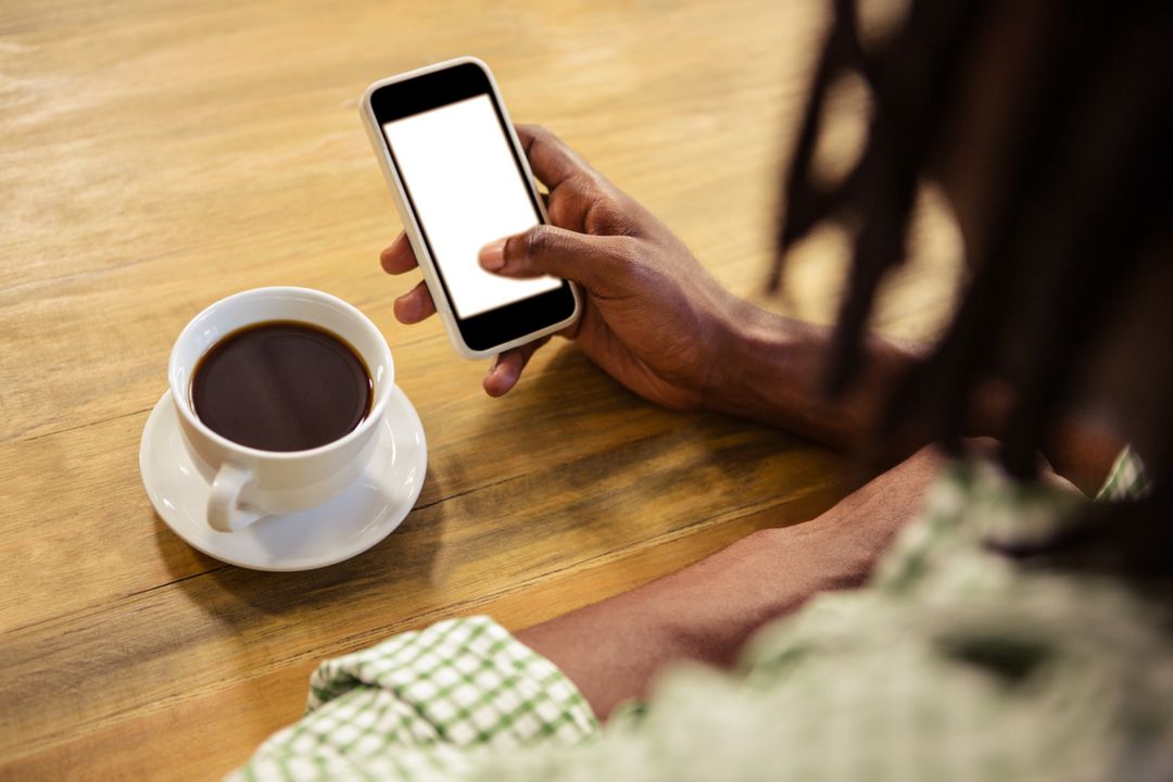 Image of a Person Holding his Phone in One Hand Next to a Cup of Coffee on a Table