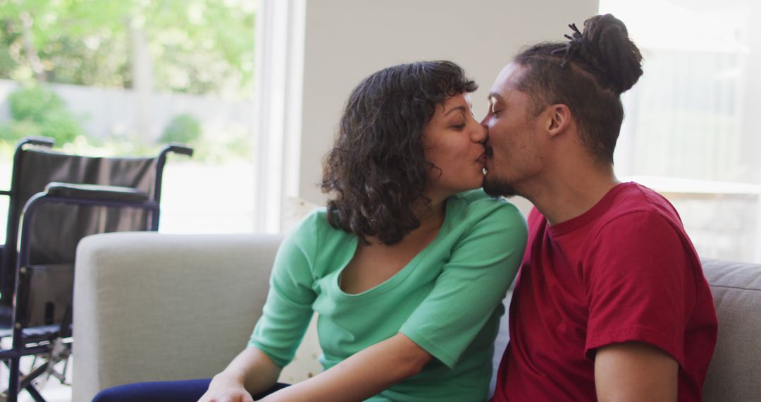 Happy biracial couple sitting on couch kissing in living room, with wheelchair in background - Free Images, Stock Photos and Pictures on Pikwizard.com