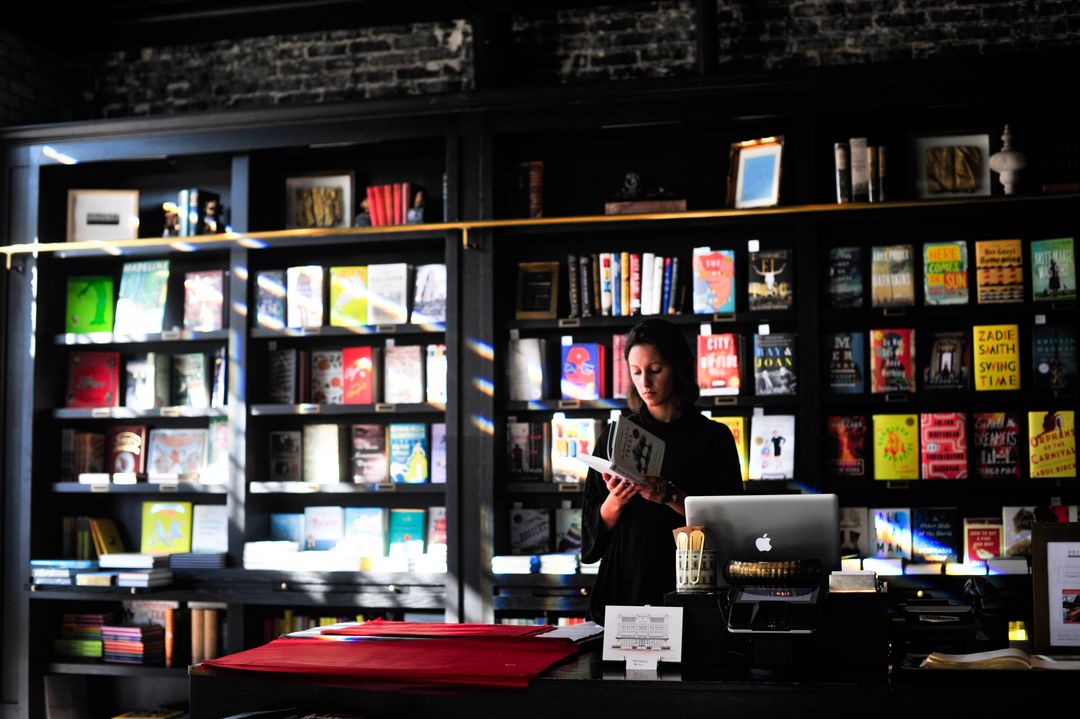 Woman standing in a book store reading a book while the sun shines through the window - Retail marketing strategy tips and tricks to drive sales and save money - Image