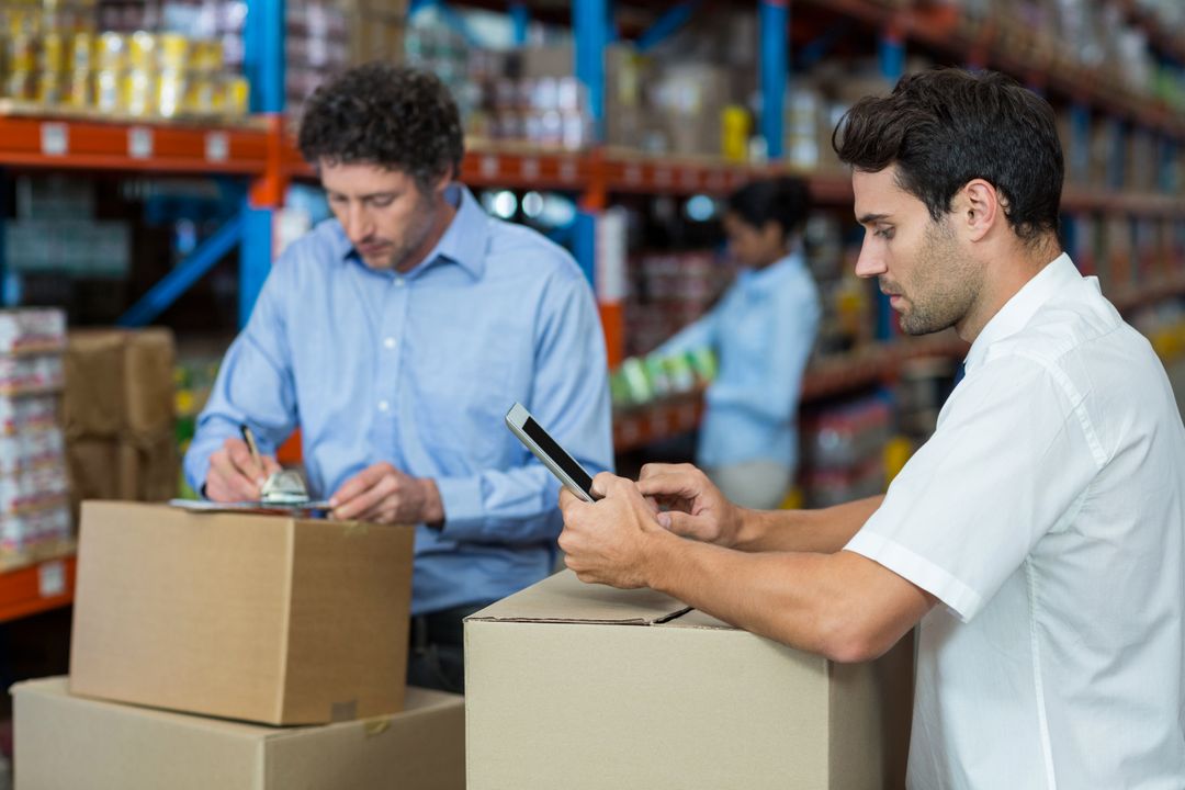 Two workers leaning on boxes in a warehouse with one looking at tablet and one writing on clipboard