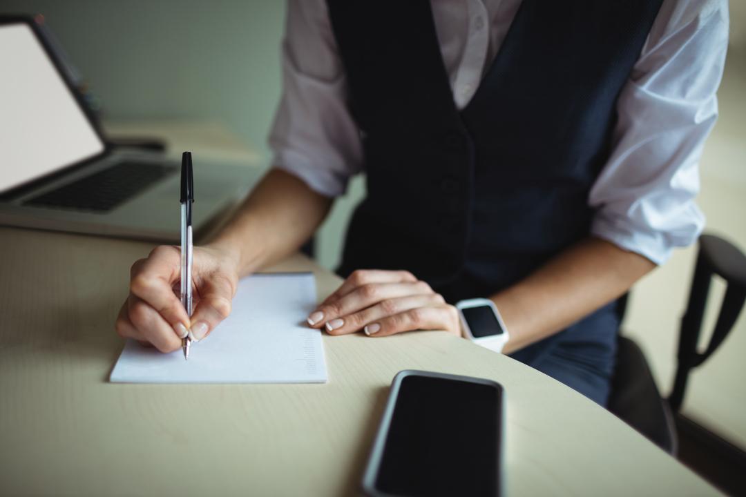 Woman writing on a notebook with a pen at a desk with laptop and phone - Essential steps to start a lifestyle blog and be successful - Image