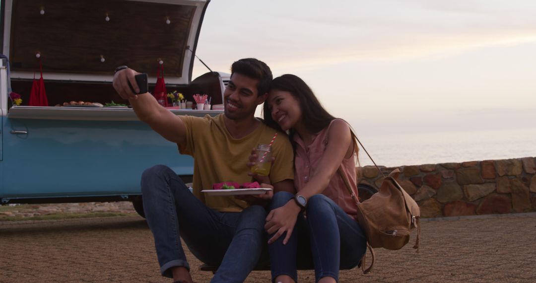 A diverse couple enjoys a sunset snack while taking a selfie