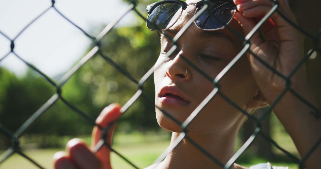 Biracial woman wearing sunglasses and holding chain-link fence at park - Free Images, Stock Photos and Pictures on Pikwizard.com