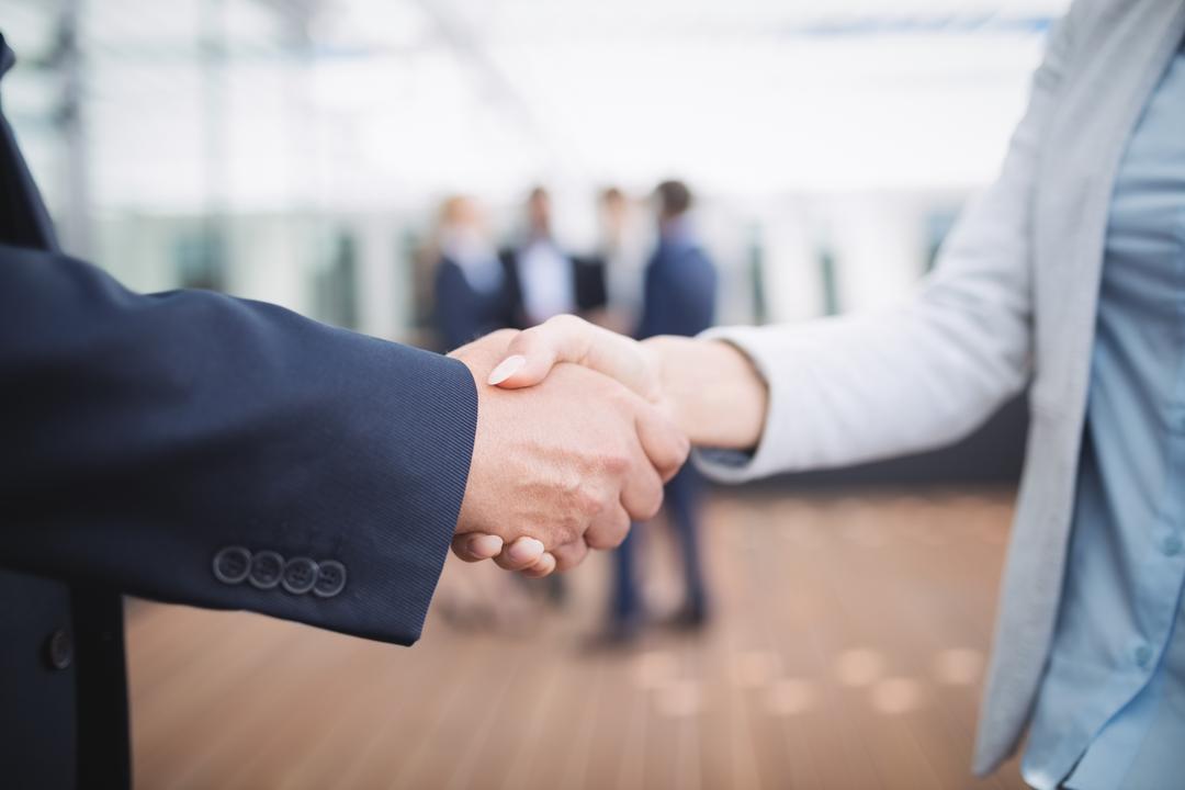 close up of two people shaking hands with workers in the background - Retail marketing strategy tips and tricks to drive sales and save money - Image