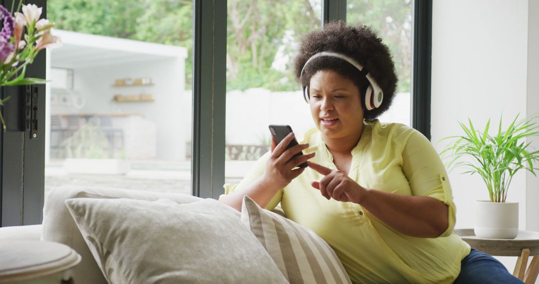 Image of happy plus size african american woman with headphones sitting on sofa with smartphone - Free Images, Stock Photos and Pictures on Pikwizard.com