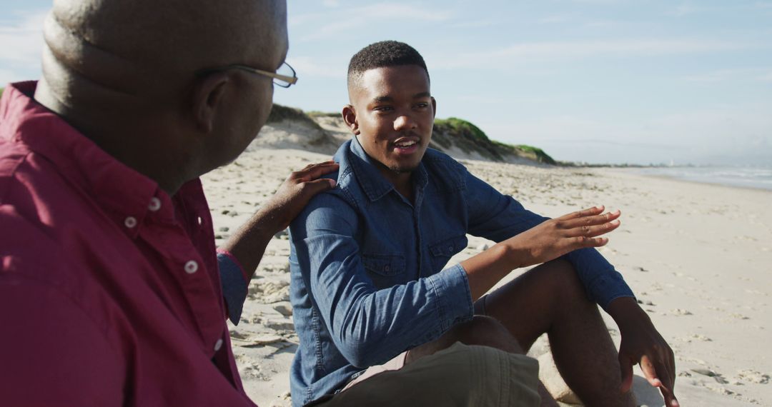 African american father sitting on beach with teenage son, putting hand on his shoulder and talking - Free Images, Stock Photos and Pictures on Pikwizard.com