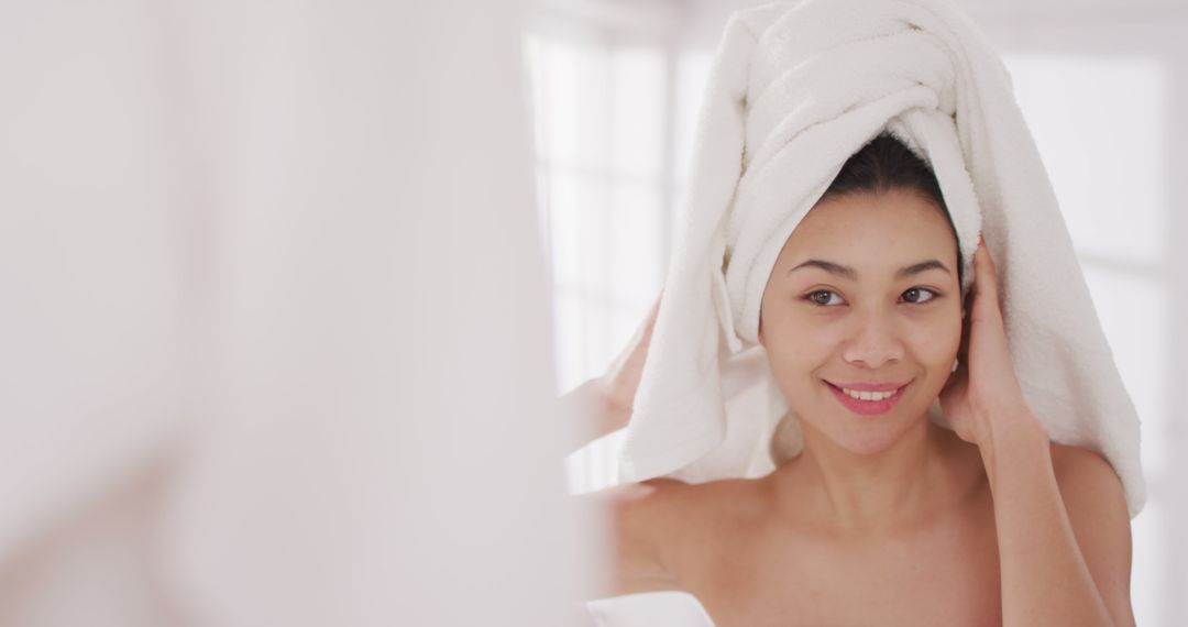 Image of portrait of smiling biracial woman with towel on hair looking in mirror in bathroom - Free Images, Stock Photos and Pictures on Pikwizard.com