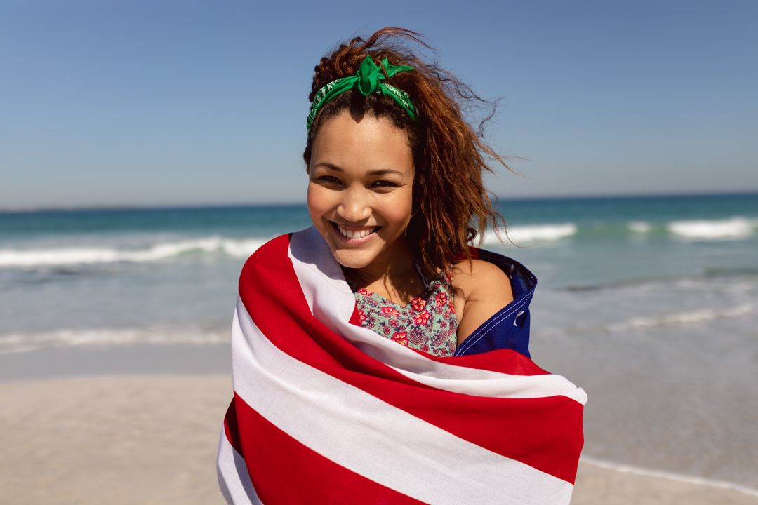 Beautiful young woman wrapped in american flag looking at camera on beach in the sunshine - Free Images, Stock Photos and Pictures on Pikwizard.com