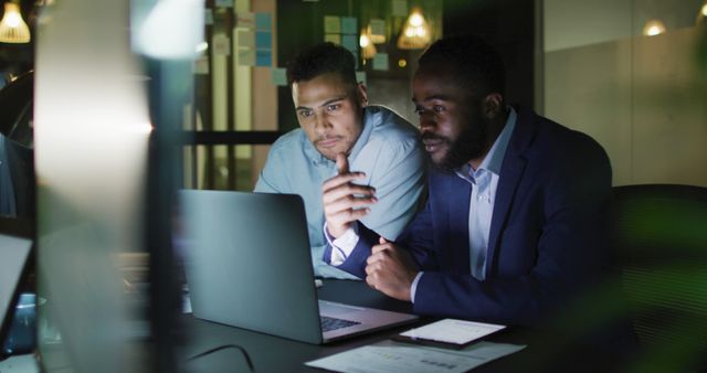 Image of two happy diverse businessmen talking and using laptop together at night in office. Business, communication, inclusivity and flexible working concept digitally generated image.