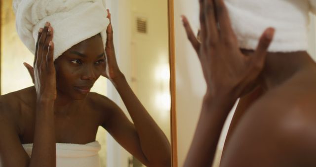 African american woman with towel touching her face and watching in mirror in bathroom - Download Free Stock Photos Pikwizard.com