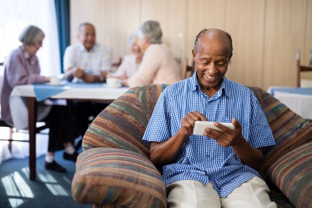 Happy senior man using phone while sitting on sofa against friends at nursing home