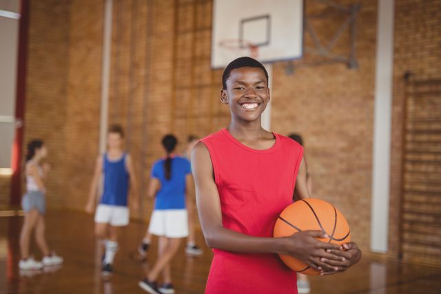 Smiling school boy holding a basketball while team playing in background - Download Free Stock Photos Pikwizard.com