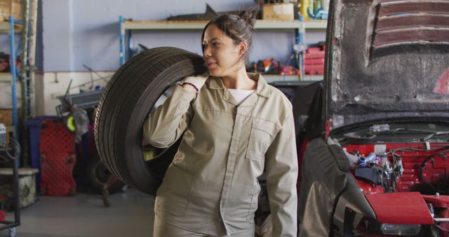 Image of happy biracial female car mechanic holding tyre. Working in car repair shop and running small feminine business concept.