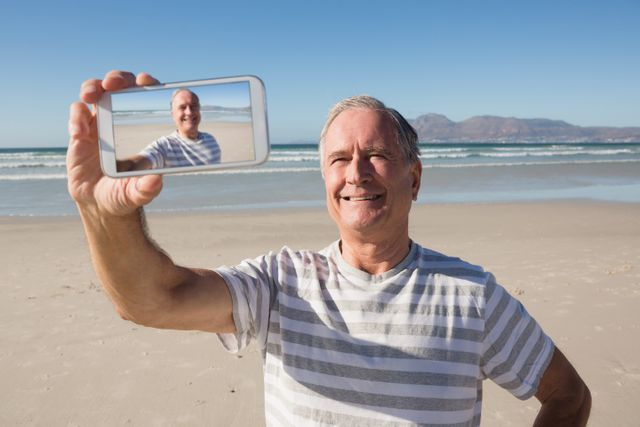 Smiling senior man holding smart phone while standing against sea at beach