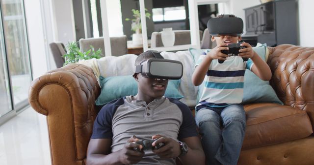 Image of happy african american father and son sitting on sofa and playing games with vr headset. Family, spending quality time together at home with technology concept.