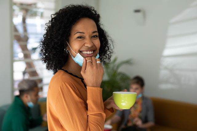 African american woman pulling her facemask down showing a smile while holding a cup of coffee. behind her are her colleagues sitting on the couch talking during their coffee break.