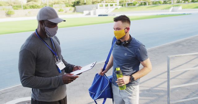 Diverse male coach and athlete wearing face mask talking before training session. professional runner training at sports stadium during coronavirus covid 19 pandemic