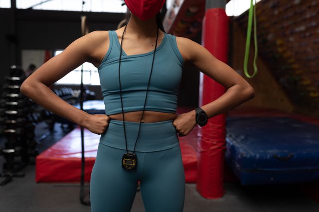 Caucasian woman wearing a stopwatch around her neck and a facemask in the gym. she is standing with her arms on her waist.