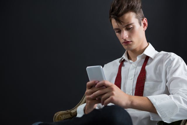 Androgynous man sitting on chair and using mobile phone against black background