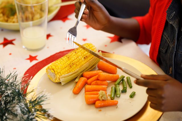 Close up of an african-american child holding a fork and a knife eating at a christmas dinner plate with a corn cob. carrots and beans beside a glass of milk.