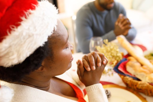 Side view of an african-american woman wearing a santa hat praying with family at a dinner table wit - Download Free Stock Photos Pikwizard.com