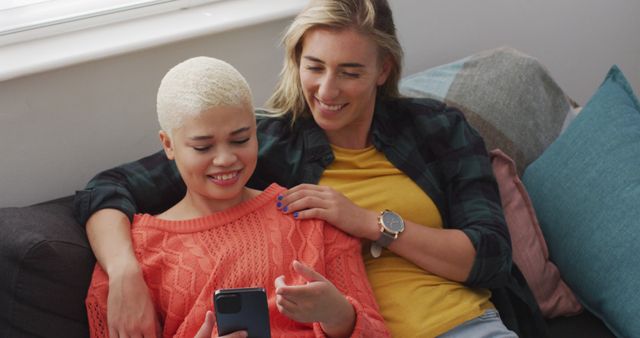 Happy diverse female couple embracing and using smartphone in living room. spending quality time at home.