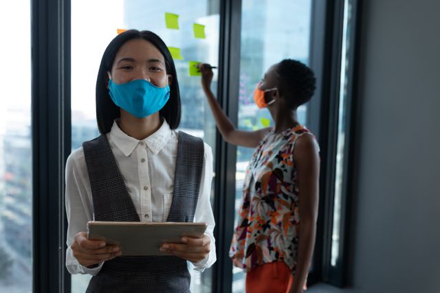 Asian woman wearing face mask using digital tablet looking at camera. with coworker in the background. health and hygiene in creative office during coronavirus covid 19 pandemic.