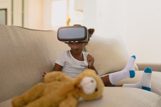 African american girl wearing vr headset and sitting on sofa with teddy bear - Download Free Stock Photos Pikwizard.com