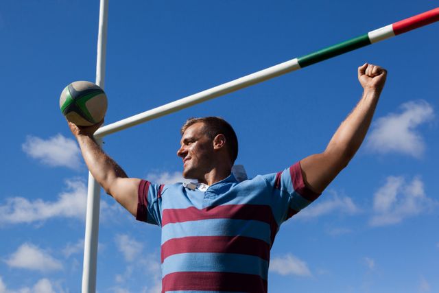 Smiling rugby player holding ball with arms raised against blue sky - Download Free Stock Photos Pikwizard.com