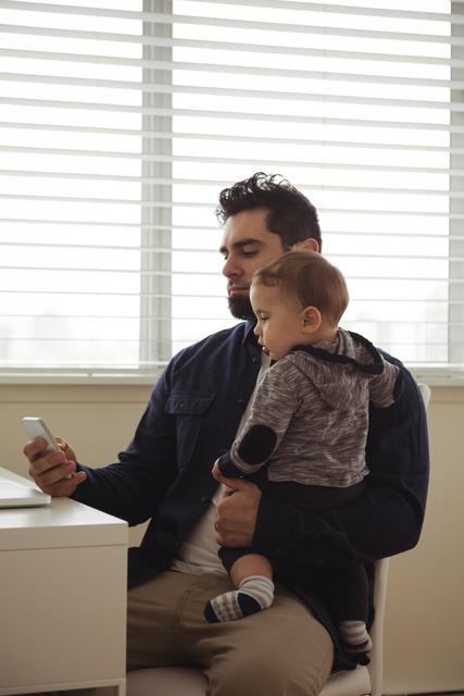 Father holding his baby while using mobile phone at desk - Download Free Stock Photos Pikwizard.com