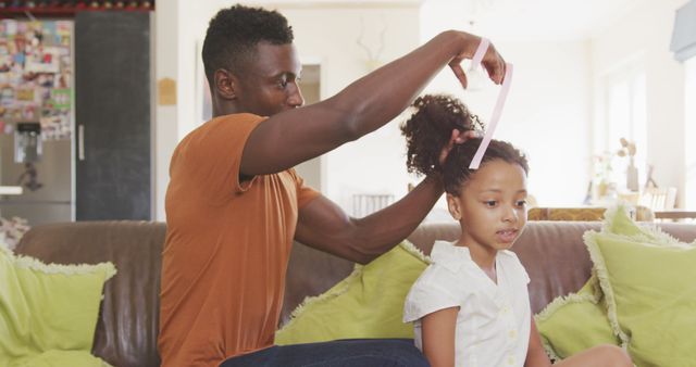 African american father tying up daughter's hair sitting on couch in living room. Fatherhood, childhood, care, togetherness and domestic life.