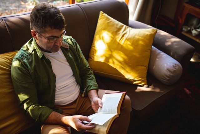 Caucasian male wearing glasses sitting on sofa and reading book. spending free time at home.
