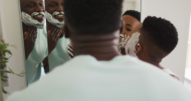 African american father and son putting shaving cream on their faces and laughing together. staying at home in self isolation during quarantine lockdown.