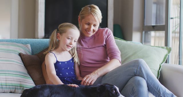 Happy caucasian mother and daughter sitting on sofa together and petting dog at home. Motherhood, childhood, care, togetherness and domestic life.