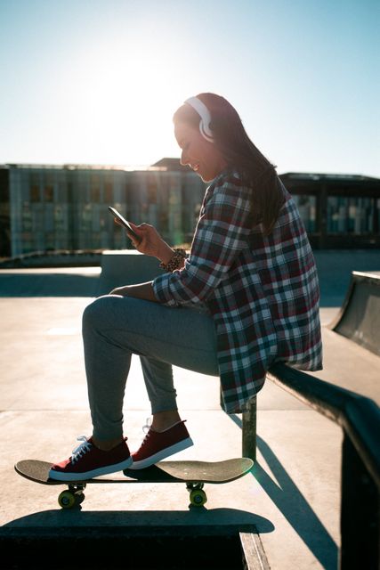 Smiling caucasian woman wearing headphones, using smartphone and sitting on handrail in the sun. hanging out at an urban skatepark in summer.