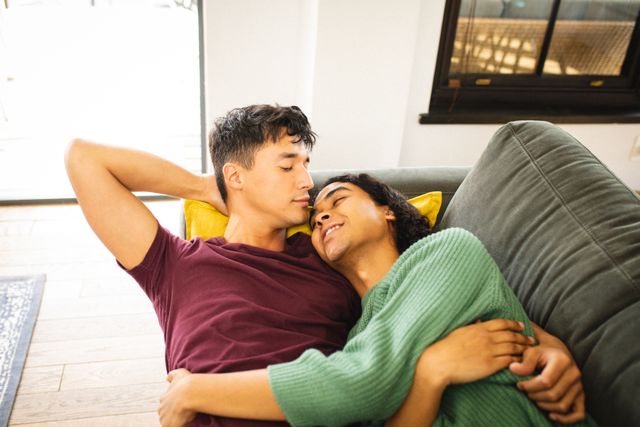 High angle view of romantic multiracial gay couple cuddling and lying on sofa in living room. Copy space, closeness, unaltered, love, togetherness, relaxing, homosexual, lifestyle and home concept.