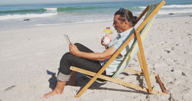 Happy senior caucasian man sitting on deckchair, using tablet and drinking cocktail on beach. Senior lifestyle, realxation, nature, communication, free time and vacation.
