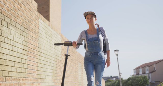Biracial woman wearing cap and overalls walking with scooter on street. Street style, modern urban lifestyle and transport.