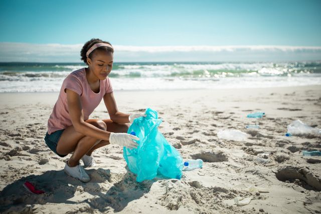 African american woman putting rubbish in refuse sack on sunny beach - Download Free Stock Photos Pikwizard.com