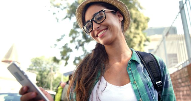 A young Caucasian woman smiles as she looks at her phone, with copy space. Dressed casually with a hat and glasses, she appears to be enjoying a sunny day outdoors.