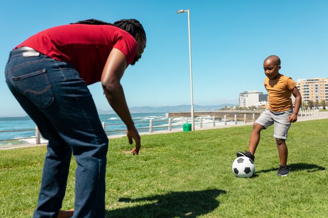African american father and son playing soccer on grass promenade at beach during sunny day. unaltered, enjoyment, family, lifestyle and togetherness concept.