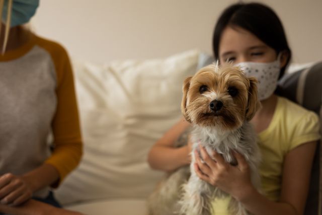 Little caucasian girl carrying a dog while sitting on the couch beside her mother at home - Download Free Stock Photos Pikwizard.com