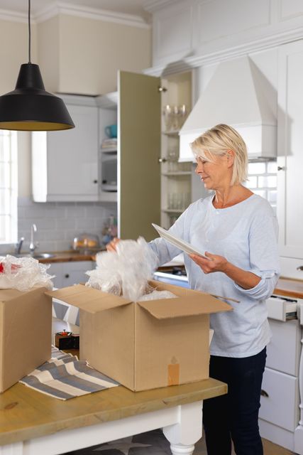 Vertical image of happy mature caucasian woman unpacking dishes in kitchen at home, copy space. Domestic life, living alone and senior lifestyle concept.