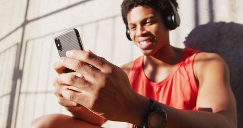 Happy african american man in city, using smartphone with wireless headphones, sitting against wall. fitness and active urban outdoor lifestyle.