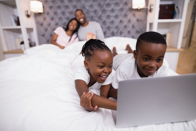 Happy siblings using laptop on bed with parents in background - Download Free Stock Photos Pikwizard.com