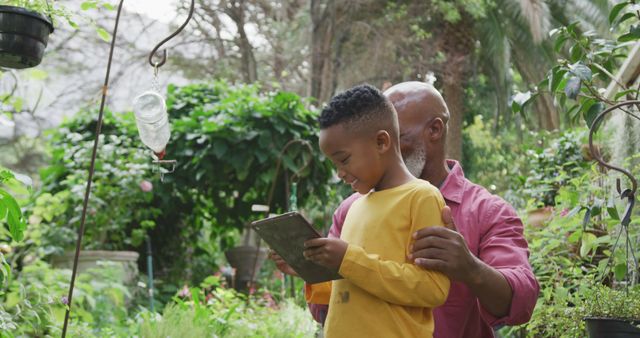 Happy senior african american man with his grandson looking at plants and using tablet in garden. Spending time outdoors, working in garden nursery.