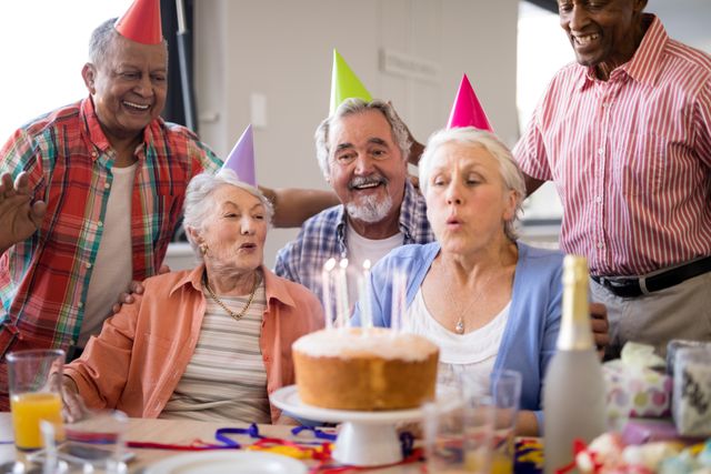 Friends looking at senior woman blowing candles on birthday cake during party in nursing home