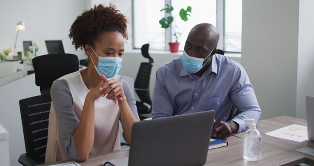 Diverse businessman and businesswoman in face masks discussing, using laptop in office. business professional working in modern office during covid 19 coronavirus pandemic.