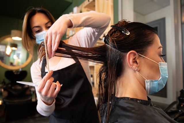 Caucasian female hairdresser working in hair salon cutting hair of female customer both wearing face masks. Health and hygiene in workplace during Coronavirus Covid 19 pandemic.