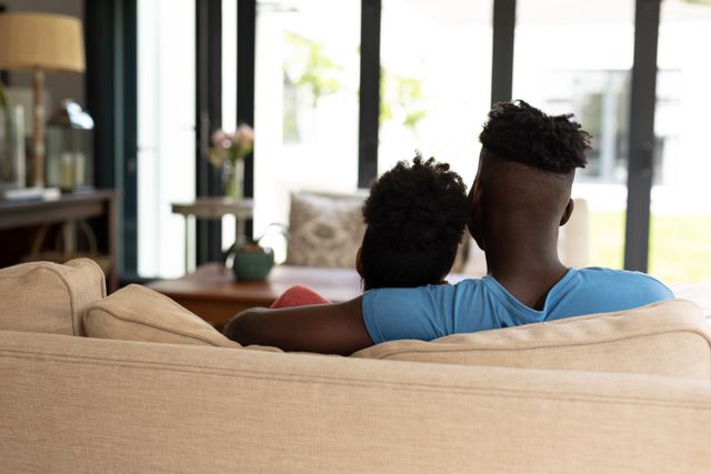 Rear view of african american young man with arm around girlfriend sitting on sofa in living room. unaltered, lifestyle, home, love and togetherness concept.